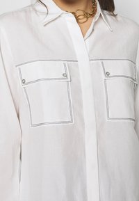White button-up shirt made of lightweight fabric, featuring two front pockets with contrasting black stitching and snap closures.