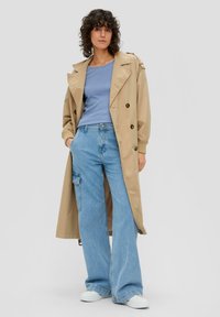 Beige trenchcoat met schouderepauletten, wijde lichtblauwe jeans en witte sneakers. Geribbelde lichtblauwe top eronder.