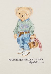 Un oso de dibujos animados con un suéter verde con la impresión "Polo", jeans azules con parches, zapatillas blancas, sosteniendo una bolsa tejida con una cinta rosa.