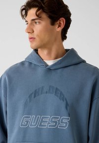 Niebieska bluza z kapturem wykonana z miękkiego materiału, wyposażona w przednią kieszeń, regulowany sznurek przy kapturze oraz nadruki z tekstem „ATHL.DERT.” i „GUESS”.