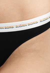 Gros plan sur un sous-vêtement noir avec une ceinture blanche présentant le logo « BJÖRN BORG » aux couleurs de l’arc-en-ciel en répétition sur un fond de peau.