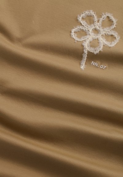 Tan tyg med mjuk textur och en broderad vit blomma som accent samt "NN. 07" i vitt. Materialet har en subtil lyster.