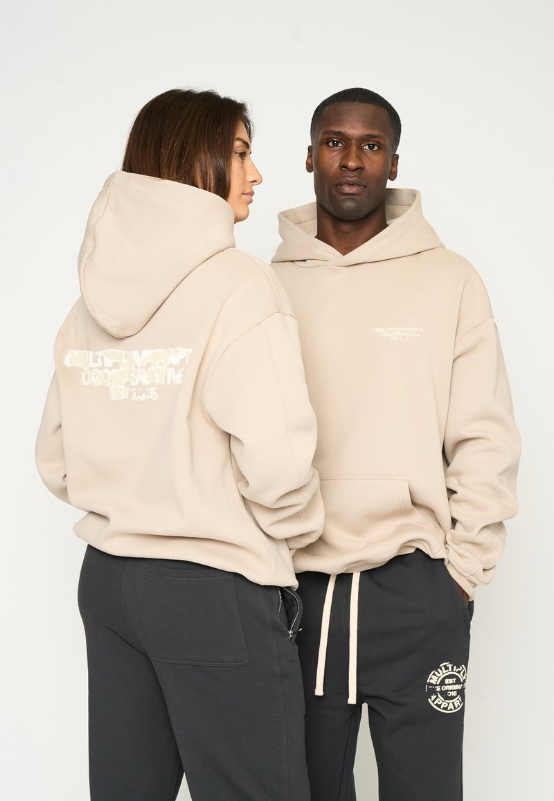 Beige hættetrøjer med et stort logo på ryggen og mindre tekst på forsiden. Kombineres med mørke sweatpants, der har et logopatch på låret.