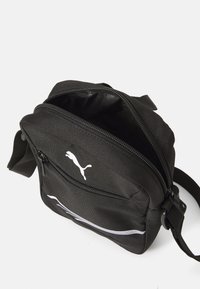 Bolso cruzado de tela negra con un bolsillo delantero con cremallera, logo de Puma blanco y una franja horizontal reflectante; cuenta con correa ajustable.