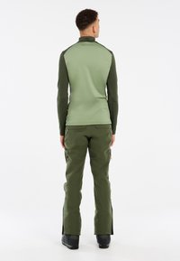 Groene longsleeve top met een opstaande kraag, gecombineerd met groene cargobroek met ritszakken en zijsplitten, en zwarte laarzen.