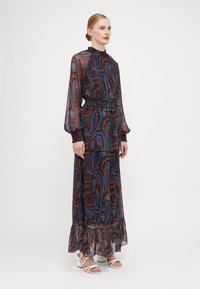 Μακρύ, μπλε φόρεμα με σχέδιο paisley σε κόκκινες και κίτρινες αποχρώσεις. Διαφανείς, φουσκωτές μανσέτες, ψηλό γιακά, ελαστική μέση και φούντωτο στρίφωμα. Λευκά παπούτσια με λουράκια.