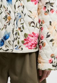 Μπουφάν λευκό με καπιτονέ ύφασμα και πολύχρωμα λουλουδάτα και πουλερικά μοτίβα, φορεμένο πάνω από σκοτεινό ελιά παντελόνι, με εμφανείς λεπτομέρειες στα μανίκια και στη μέση.