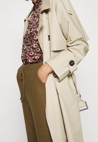 Trench-coat beige à coupe décontractée, doté de grands boutons et d'une ceinture. En dessous, un chemisier à motif floral et un pantalon vert olive.