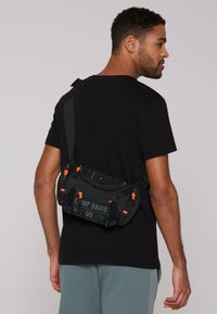 Black canvas crossbody bag with orange accents, logo, durable zippers, and textured pattern, worn over the shoulder.