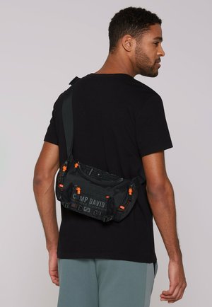 Schwarze Canvas-Crossbody-Tasche mit orangefarbenen Akzenten, Logo, strapazierfähigen Reißverschlüssen und strukturierter Oberfläche, über der Schulter getragen.