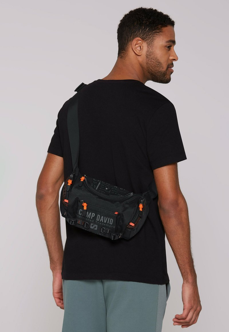 Black canvas crossbody bag with orange accents, logo, durable zippers, and textured pattern, worn over the shoulder.