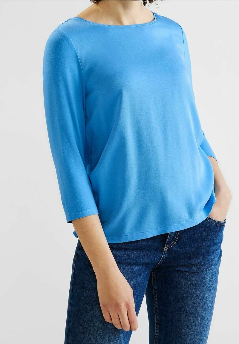 Sininen, kevyt paita, jossa on pyöreä pääntie ja kolmen neljänneksen hihat. Paita on sileä ja rento istuvuus, yhdistettynä farkkuihin.