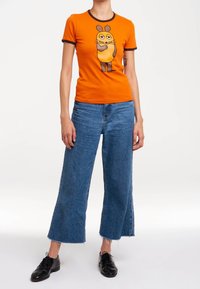 T-shirt orange à manches courtes et encolure bordée de marron, avec un motif de souris de dessin animé ; associé à un jean bleu à jambes larges et des chaussures noires.