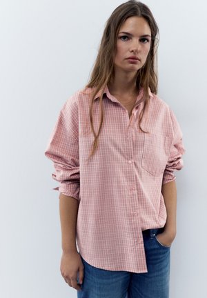 Jeune femme aux longs cheveux portant une chemise rose à carreaux ample et un jean bleu, debout avec une main dans la poche devant un fond uni.