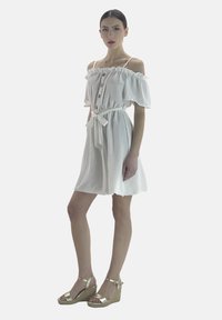 Vestido blanco sin mangas con volantes en las mangas, detalle de botones y cintura con lazo. Llevado con sandalias de cuña doradas sobre un fondo liso.