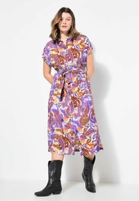 Bunt gemustertes, knielanges Kleid mit kurzen Ärmeln, einem gebundenen Taillenzug und einer Knopfleiste. Kombiniert mit schwarzen Cowboy-Stiefeln.