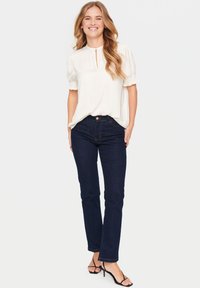 Blusa bianca a maniche corte con dettaglio a chiave sul davanti, abbinata a jeans blu scuro a gamba dritta e sandali neri con cinturini. Texture liscia.
