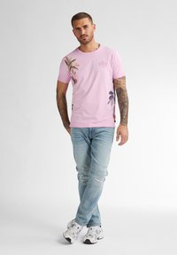 Camiseta de algodón rosa con estampados de palmeras, cuello redondo, mangas cortas y un sutil logo; combinada con jeans ligeros rasgados de color azul y zapatillas blancas.