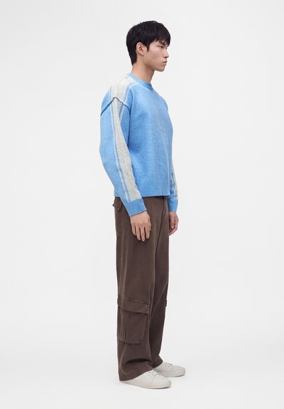 Jeune homme debout de profil, portant un pull bleu et gris, un pantalon cargo marron et des baskets blanches sur un fond blanc uni.