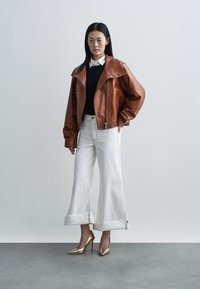 Femme portant une veste en cuir marron, un pull noir, une chemise blanche à col, un jean blanc large avec des revers, et des talons hauts dorés, se tenant devant un fond uni.
