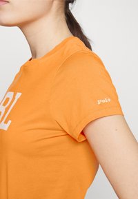 Orange bomullströja med en vit grafik "GIRL" på framsidan och en liten "Polo"-logotyp på vänster ärm. Kort ärm och rund halsringning.