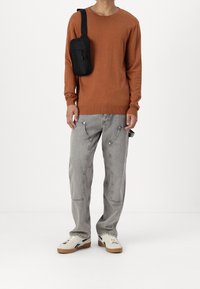 Osoba w brązowej swetrze w kolorze rdzy, szarych dżinsach z dużymi przednimi kieszeniami, białych trampkach i czarnej torebce na ramię.