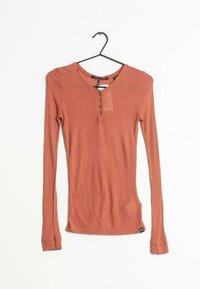 Långärmad ribbad topp i terrakotta med rund halsringning, fyra knappar och en liten etikett nära nederkanten.