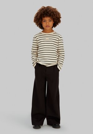 Koszula w kremowym kolorze z czarnymi paskami i długimi rękawami, zestawiona z szerokimi czarnymi spodniami. Koszula ma małą naszywkę z logo na piersi.