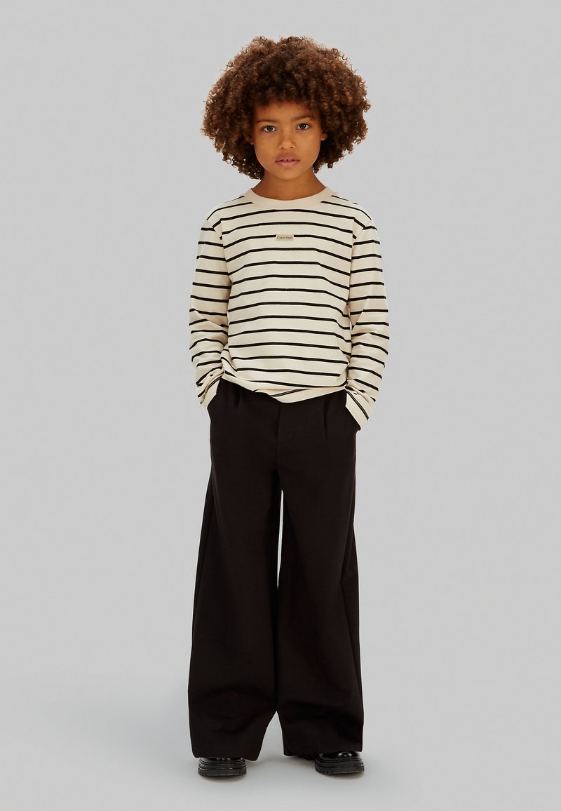 Chemise à manches longues rayée crème avec des rayures noires, associée à un pantalon large noir. La chemise est ornée d'une petite étiquette logo au niveau de la poitrine.