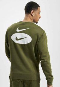 Alyvos spalvos džemperis ilgomis rankovėmis, su dideliu baltu Nike logotipu ir akcentiniu grafinu nugaroje. Minkšta tekstūra, apvalios iškirptės dizainas.