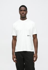 Vit bomullst-shirt med rund halsringning, korta ärmar och en svart logga tryckt på vänster sida. Enkel, avslappnad passform.