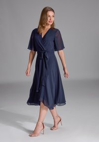 Sukunya biru navy dengan lengan pendek transparan, pinggang terikat, dan hem yang mengalir. Dipasangkan dengan sandal hak tinggi metalik yang bertali.
