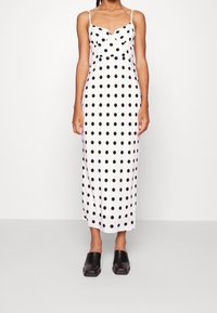 Kvinna som bär en vit klänning med svarta prickar och svarta slip-on skor, som står mot en enkel vit bakgrund.