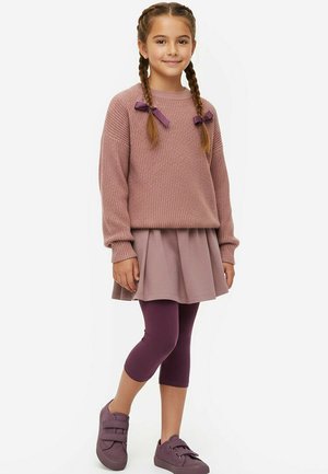 Młoda dziewczyna z warkoczami, ubrana w bladoróżowy sweter, pasującą spódnicę, ciemnofioletowe legginsy i fioletowe tenisówki, stojąca na białym tle.