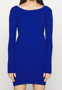 Pletené šaty v živé modré barvě, s širokým výstřihem, dlouhými rukávy, přiléhavým střihem a žebrovanou texturou. Spodní lem končí těsně nad středem stehna.