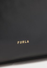 Materiale in pelle nera con una texture liscia, caratterizzato dal nome del marchio "FURLA" in rilievo dorato ben visibile.