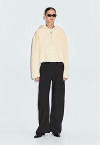 Felpa a maglia crema con mezza zip, vestibilità comoda, abbinata a pantaloni neri a gamba larga e scarpe nere con punta.