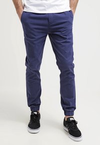 Man in een marineblauwe joggerbroek met elastische boorden en zwarte sneakers, staande met handen in de zakken tegen een witte achtergrond.