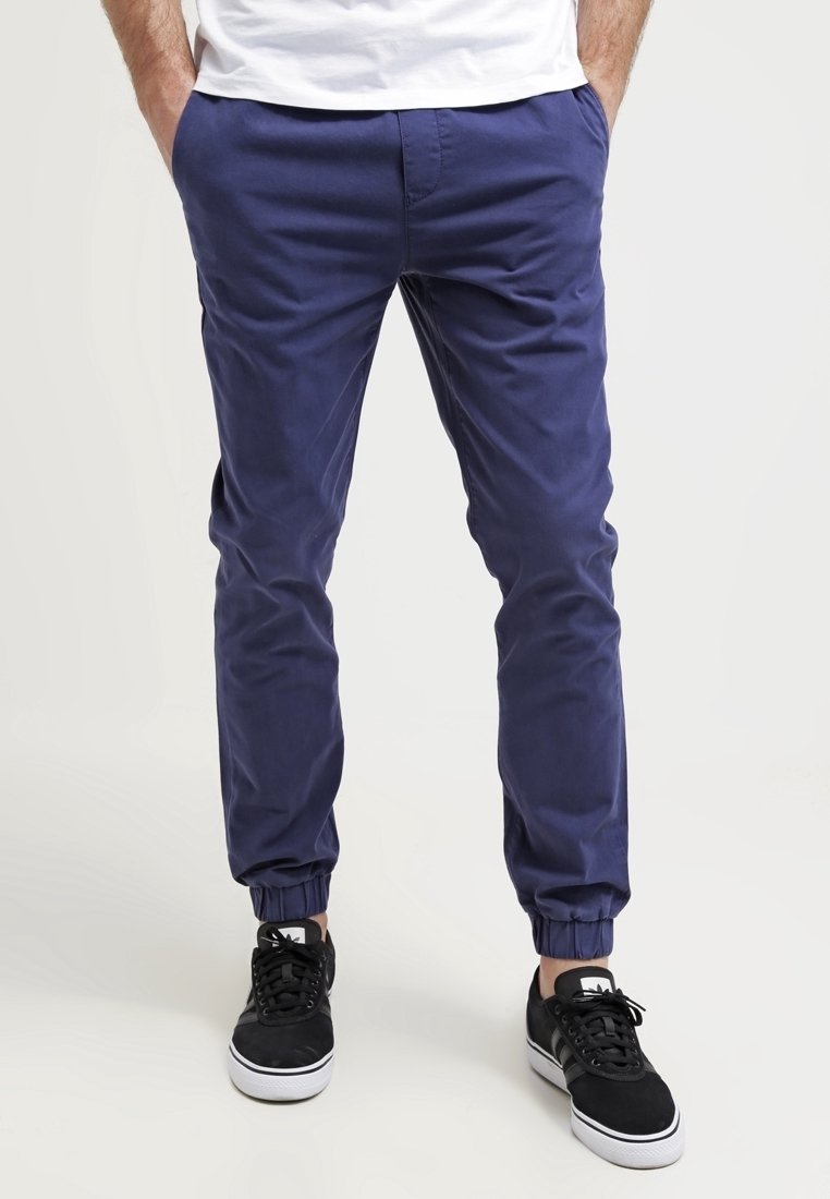 Man in een marineblauwe joggerbroek met elastische boorden en zwarte sneakers, staande met handen in de zakken tegen een witte achtergrond.