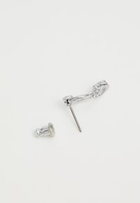 Srebrna kovinska uhan z ukrivljenim dizajnom in prozornim kamnom kot poudarkom ter ločenim zapiranjem na žebljiček. Gladka tekstura z odsevno površino.