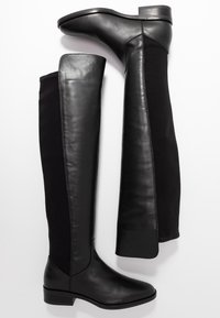 Bottes noires hauteur cuisse en cuir lisse avec des panneaux en tissu élastique, dotées d’un petit talon bloc et d’un design épuré.