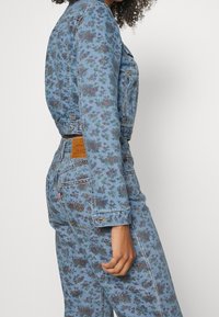 Denim jumpsuit i lyseblå med blomstermønster. Har et tætsiddende design, lange ærmer og synlige lomme detaljer.