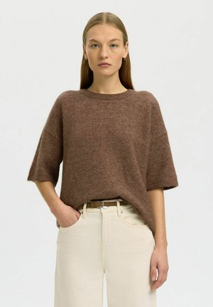 Mladá žena s rovnými blond vlasmi nosí voľný hnedý sveter a krémové vysoký pás rifle, stojí s jednou rukou v vrecku.