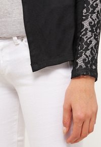 Main reposant à côté d’un pantalon blanc, portant un haut à manches longues noir avec un poignet à motif en dentelle et un pull gris en dessous.