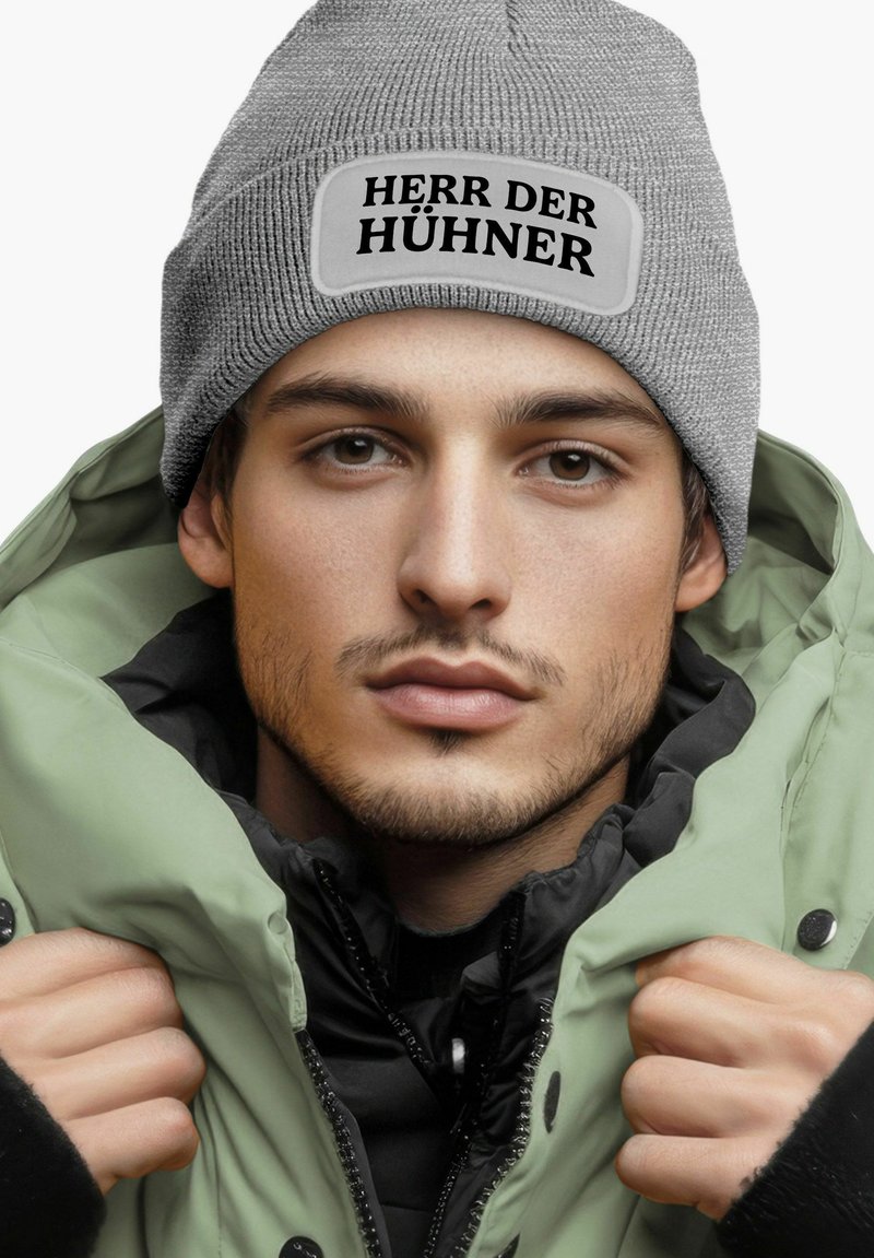 Gray knit beanie with "HERR DER HÜHNER" patch, worn with a light green puffy jacket and black inner layer, showcasing textured fabric and stitching.