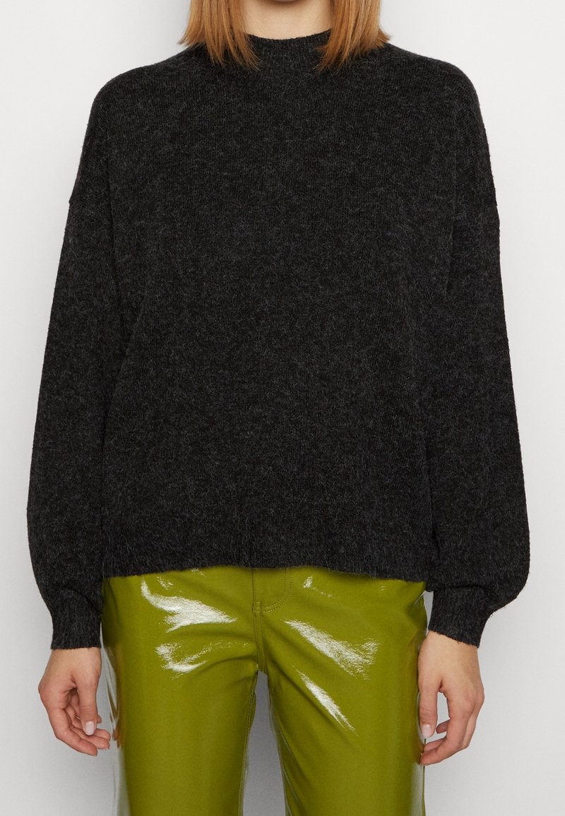 Personne portant un pull à manches longues noir et pelucheux, associé à un pantalon vert olive brillant, les mains détendues le long du corps.