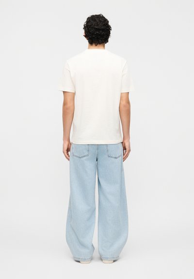 Άνδρας με σγουρά σκούρα μαλλιά, φοράει απλό λευκό κοντομάνικο μπλουζάκι και άνετο ανοιχτό μπλε τζιν, στέκεται με την πλάτη γυρισμένη σε λευκό φόντο.