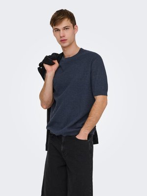 Jonge man die een marineblauw, gebreid T-shirt met korte mouwen en een zwarte broek draagt, met een zwart jack over zijn schouder, tegen een effen achtergrond.