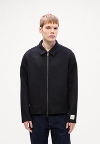 Veste noire zippée avec un col, présentant une texture lisse et une étiquette sur la manche. Associée à un jean en denim.