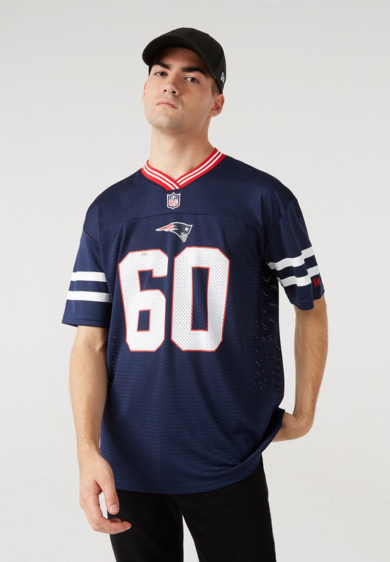 Mladý muž nosící tmavě modrý dres New England Patriots s číslem 60 a černou baseballovou čepicí, stojící před šedým pozadím.